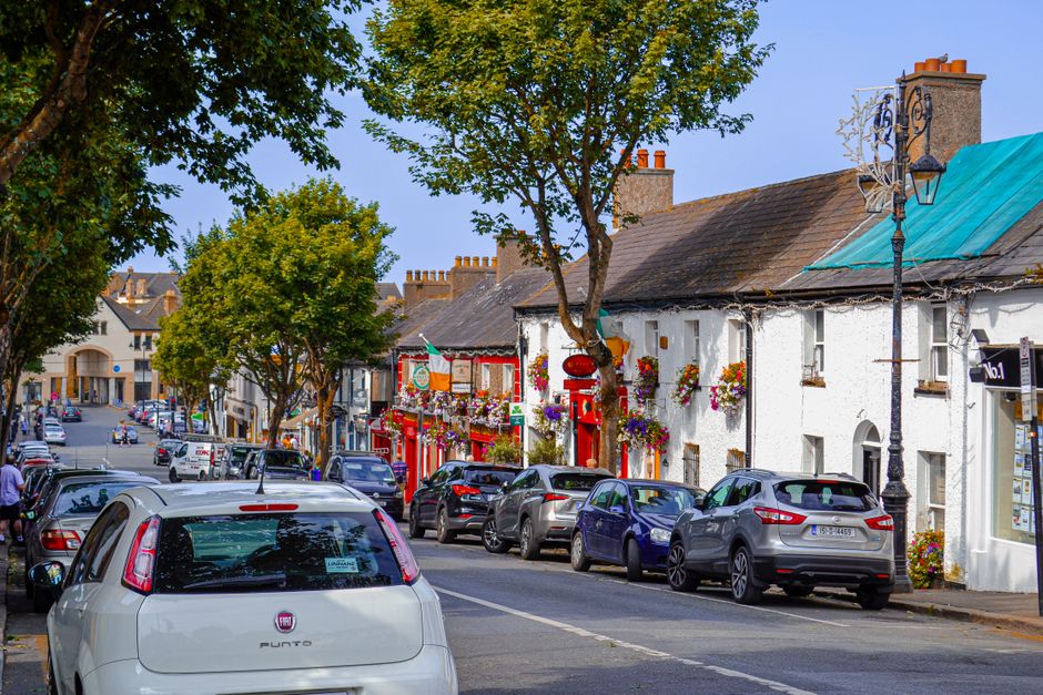 A row of charming white and colorful buildings along a busy street in Longford town center, decorated with vibrant window boxes filled with multicolored flowers. Parked cars line the street, with a wh