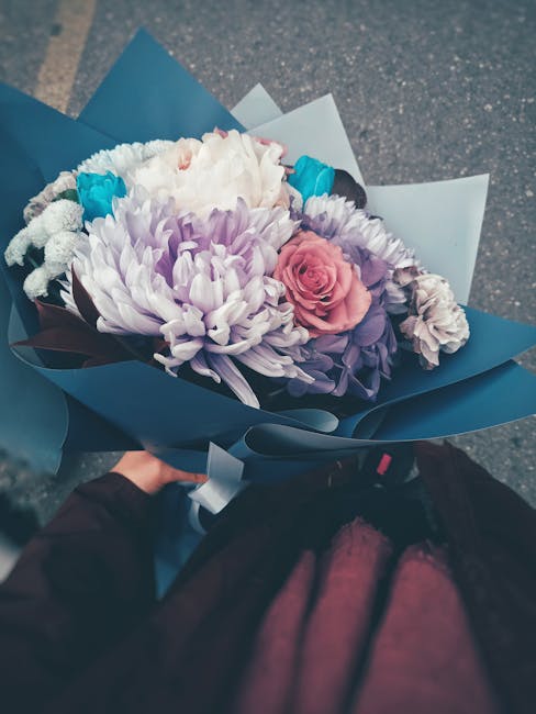 A bouquet of fresh flowers held by a person in a dark jacket, consisting of large white, purple, and pink chrysanthemums, accented with smaller blue and white blooms, wrapped in elegant blue and white