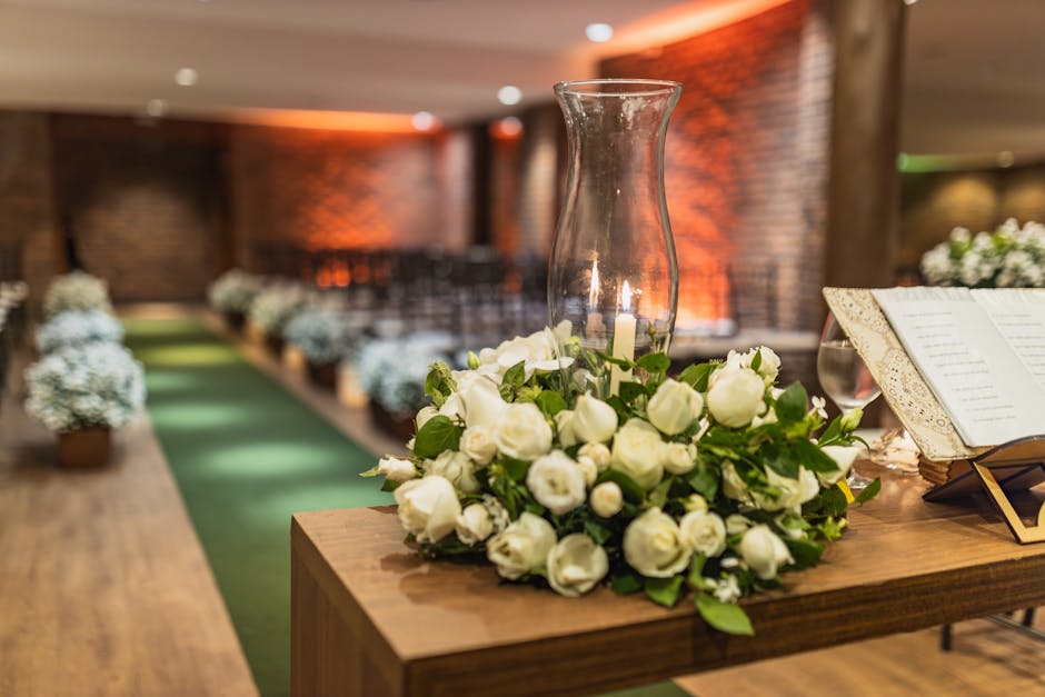 A floral arrangement featuring a cluster of fresh white roses and green foliage, placed around a glass hurricane vase on a wooden table. The arrangement appears to be part of a wedding or ceremony dec