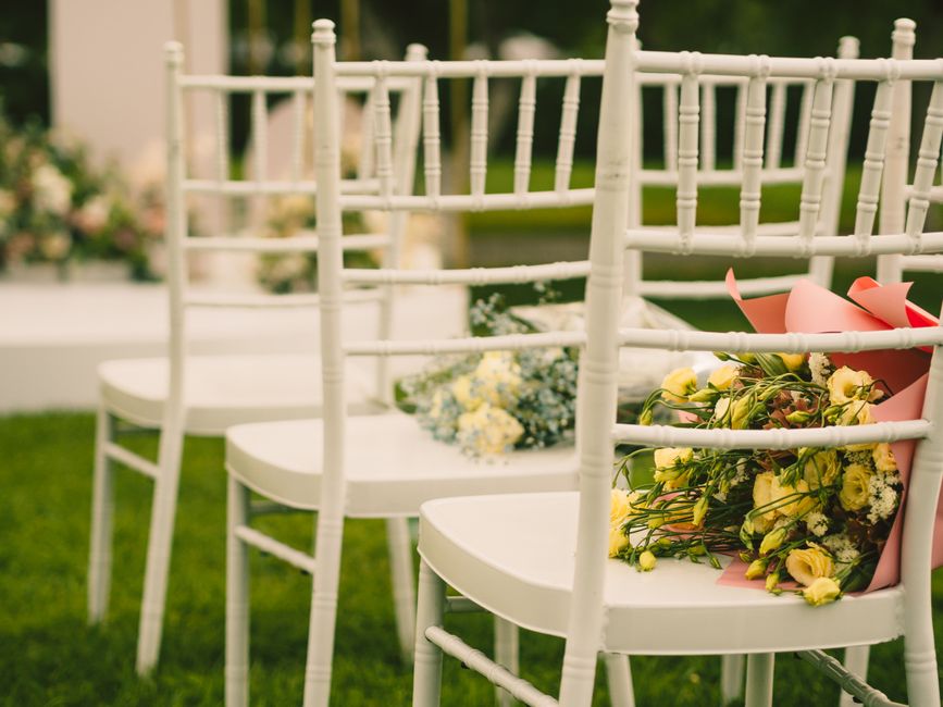 A row of white wooden chairs arranged outdoors on a grassy area, each adorned with floral arrangements. The nearest chair features a bouquet with yellow and white flowers, wrapped in pink paper, with 