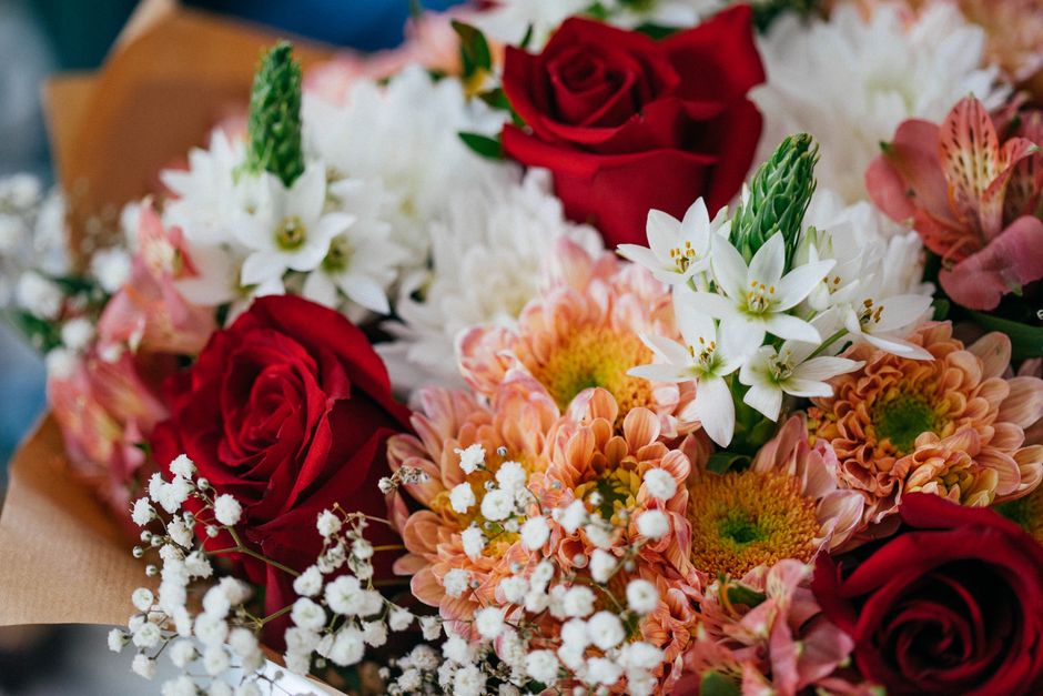 A vibrant floral bouquet arranged with deep red roses, peach and orange chrysanthemums, white star-shaped lilies, and delicate white gypsophila, all wrapped in brown craft paper. The flowers appear fr