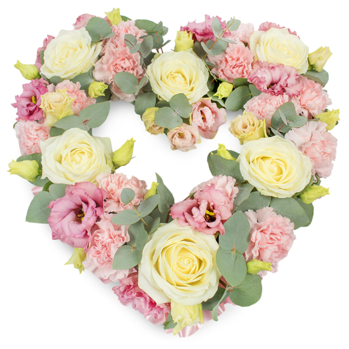 This image shows a meticulously crafted heart-shaped tribute, its generous proportions and dense florals forming a complete, unbroken heart against a stark white background. The arrangement is lavish yet composed, with every section filled to create a feeling of fullness and comfort. Large, creamy roses in shades of ivory and pale butter yellow are scattered evenly throughout the heart, each rose displaying a neat spiral of petals that open gently outwards, symbolising purity and enduring love. Nestled closely between them are numerous clusters of pastel pink carnations, their soft, frilled petals catching the light and adding both volume and a gentle, tactile feeling, almost like layers of fine fabric. Adding depth and contrast, richer magenta and mauve lisianthus blooms appear here and there, their more open, softly ruffled cups hinting at a slightly wilder, garden-grown charm. Cool, silvery-green eucalyptus foliage winds gracefully through the flowers, its round leaves overlapping and curving to echo the shape of the heart, while also introducing a calm, aromatic greenness. Tiny yellowish-green buds punctuate the design, suggesting freshness and a sense of continuation beyond loss. The overall colour palette-creams, blush pinks, mauves, and soft greens-feels soothing and dignified, ideal for a farewell where warmth and serenity are desired. From the perspective of a Longford florist, this is the kind of heart wreath a family might send to a funeral at a local chapel or crematorium, or have delivered to a home near the village centre, as a refined, heartfelt emblem of remembrance and gratitude for a life deeply loved.