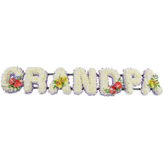 This image shows a beautifully crafted funeral flower tribute spelling "GRANDPA" in large, three-dimensional block letters, arranged in a straight horizontal line. Every letter is formed entirely from densely packed, crisp white chrysanthemums, each bloom facing outward so that the petals create an even, plush surface that feels tranquil and pure. The consistent height and width of the letters give them a solid, reassuring presence, set firmly on dark green support bars that disappear into the background while maintaining the structure. Along the base of each letter, a band of pale purple ribbon adds a subtle glow of colour, the satin edging catching faint reflections and lending the design a refined, finished look. Against the plain white backdrop, the flowers appear extra bright, as though just arranged in a cool workshop near the heart of Longford. Four thoughtfully placed sprays of accent flowers break up the white and infuse the tribute with warmth and character. On the far left "G" and the central "D," vibrant red anthuriums with glossy, heart-shaped spathes and prominent yellow spadices are grouped with deep green foliage and tiny hints of filler blooms, creating pockets of rich, emotional colour. On the first and final "A," clusters of yellow-green orchids, likely cymbidiums, arc gracefully from beds of greenery, their fresh tones suggesting renewal and enduring love. Together, the textures, hues, and clean lines express deep respect and affection for a grandfather, making this a fitting focal piece for a funeral or memorial service anywhere in the Longford community, from quiet village chapels to more modern local venues.